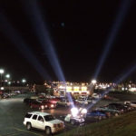 Nighttime parking lot with searchlights and cars.